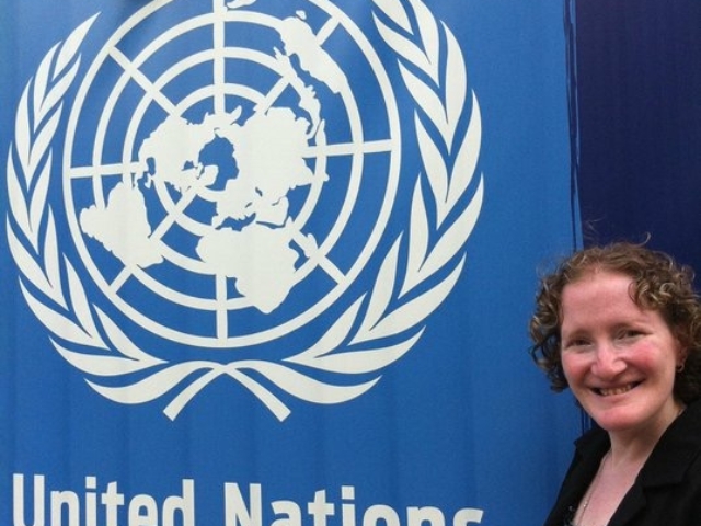 Rhonda standing next to a large blue United Nations sign. Symbol the world with olive branches.