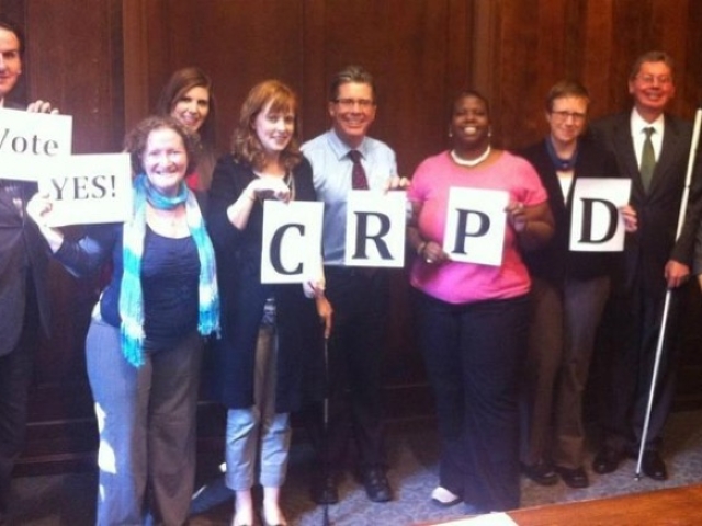 A group of 9 CRPD advocates with disabilities standing in a line holding oup papers which spell out Vote YES CRPD!