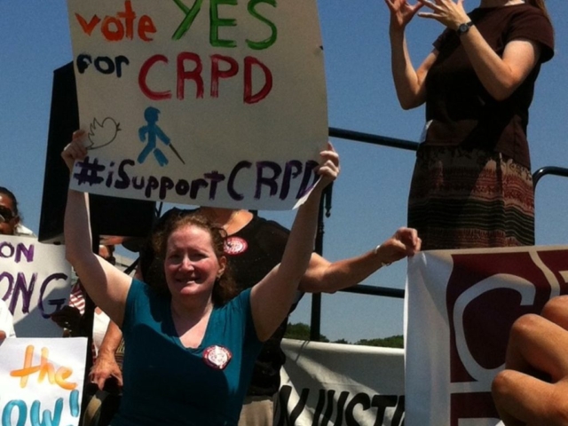 Rhonda sitting in a wheelchair holding up a sign saying "vote YES for CRPD #isupportCRPD." A sign language interpreter is interpreting behind her.