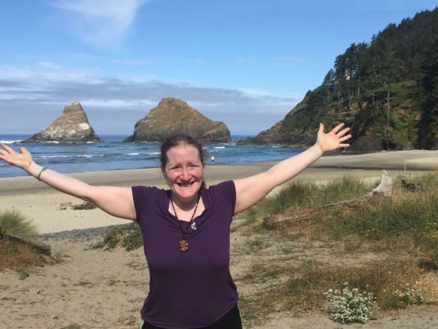Rhonda with her arms raised in the air with the pacific ocean and large rocks behind her.