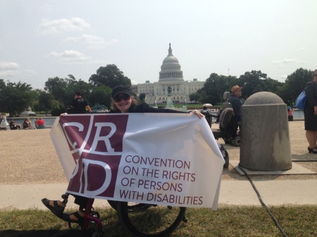 Rhonda sitting in a wheelchair, holding a big banner with the words of CRPD on it, in front of US Capitol