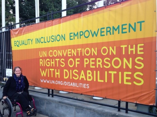 Rhonda using a wheelchair in front of a large sign at the US that says "Equality- Inclusion - Empowerment. UN Convention on the Rights of Persons with Disabilities. www.un.org/disabilities