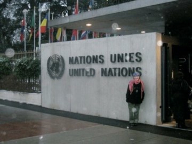 Rhonda in front of a large United Nations building and sign, with global flags in the distance.